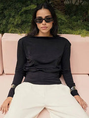 Woman wearing a black long-sleeve top and white pants sitting on a pink couch with greenery in the background.