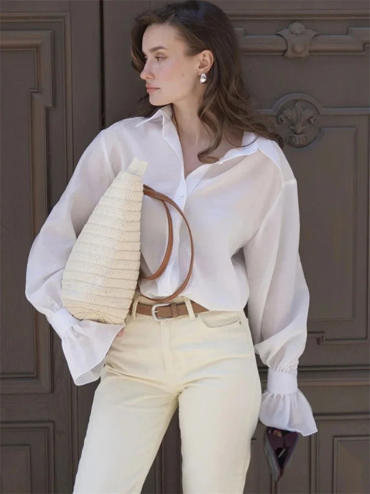 Woman wearing a white blouse and light-colored pants holding a woven bag against a wooden door.