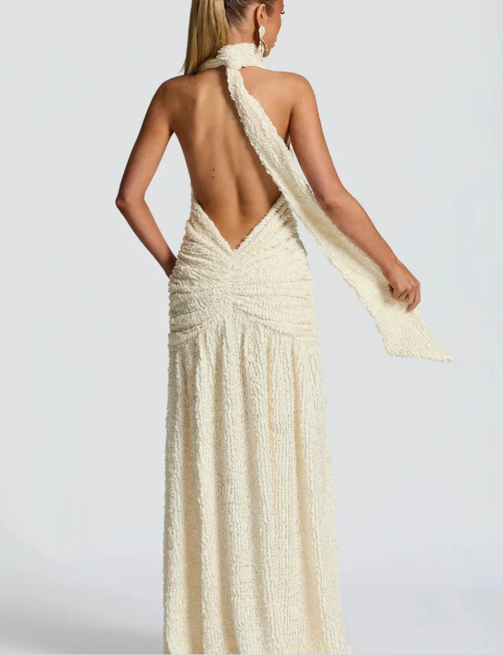 Woman wearing a long, elegant dress with a backless design and a flowing fabric over one shoulder on a plain background.