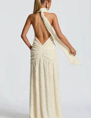 Woman wearing a long, elegant dress with a backless design and a flowing fabric over one shoulder on a plain background.