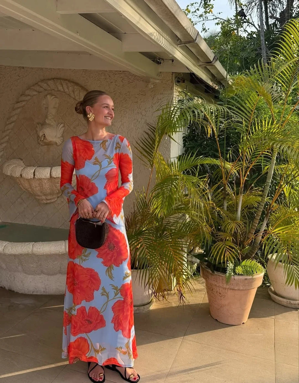 Woman in a floral dress standing outdoors near plants and a fountain.