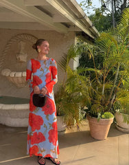 Woman in a floral dress standing outdoors near plants and a fountain.