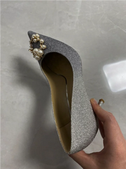 Silver glittery high heel shoe with pearl embellishments held by a hand on a gray background