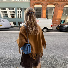 Person wearing a brown fringed jacket walking on a street with buildings and cars in the background.