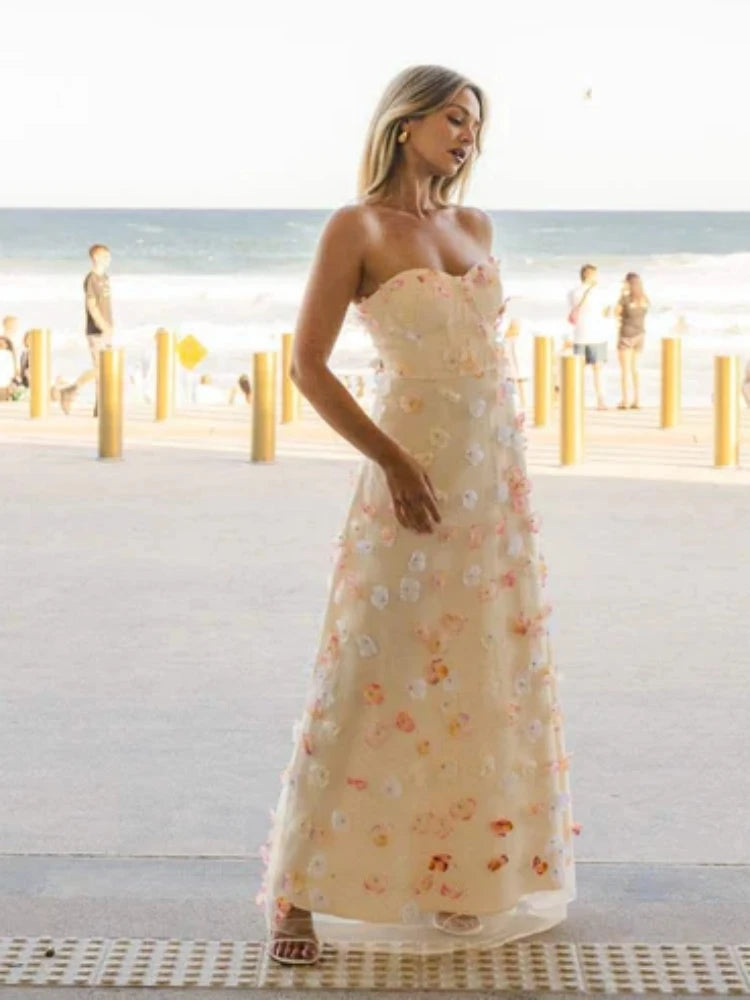 Woman in a strapless floral dress standing on a beach with people in the background.