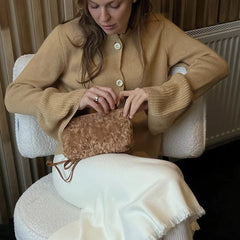 Woman sitting on a chair wearing a beige cardigan and white pants, holding a brown textured bag.