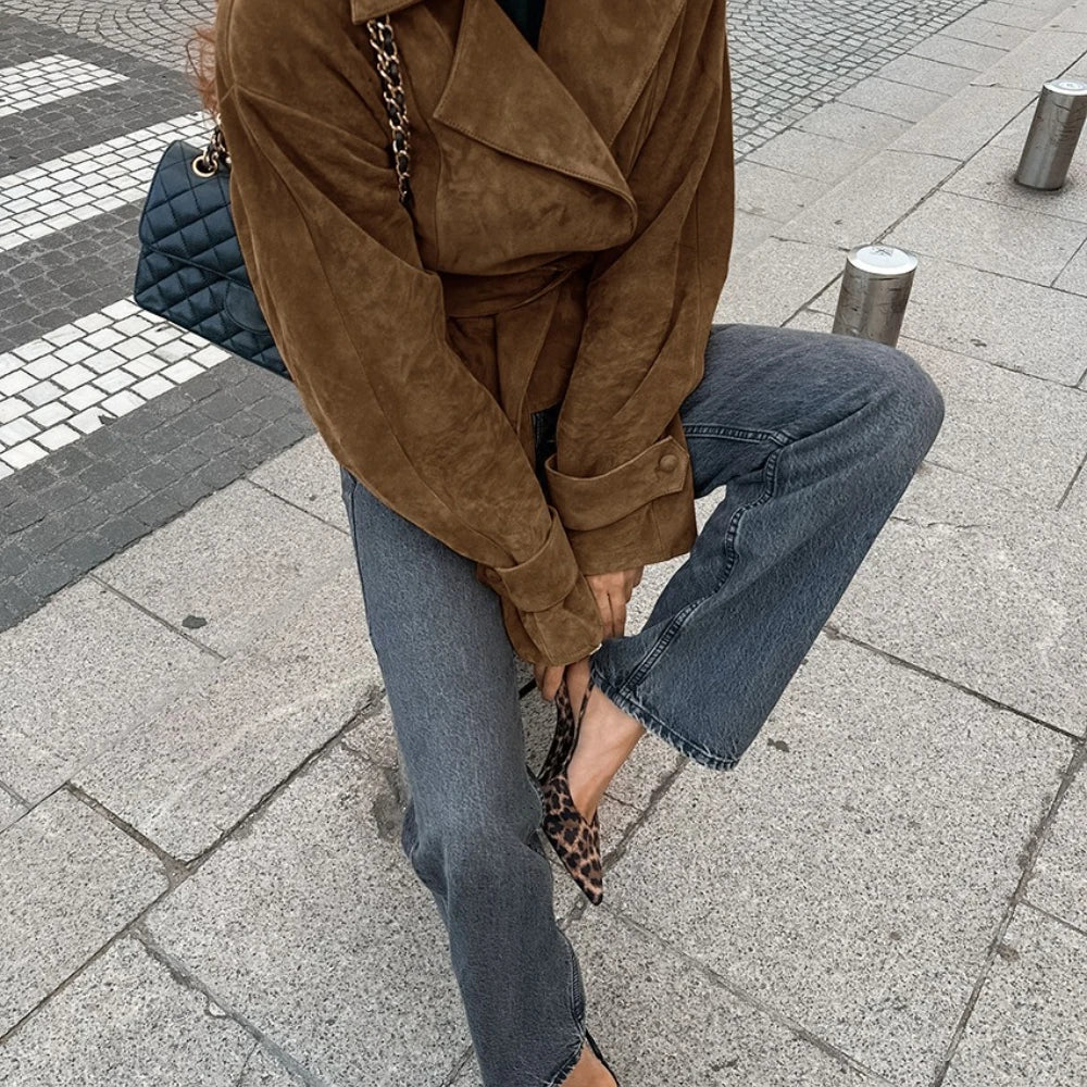 Person wearing a brown suede jacket and blue jeans on a city street.