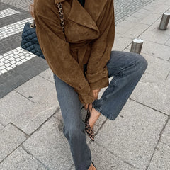 Person wearing a brown suede jacket and blue jeans on a city street.