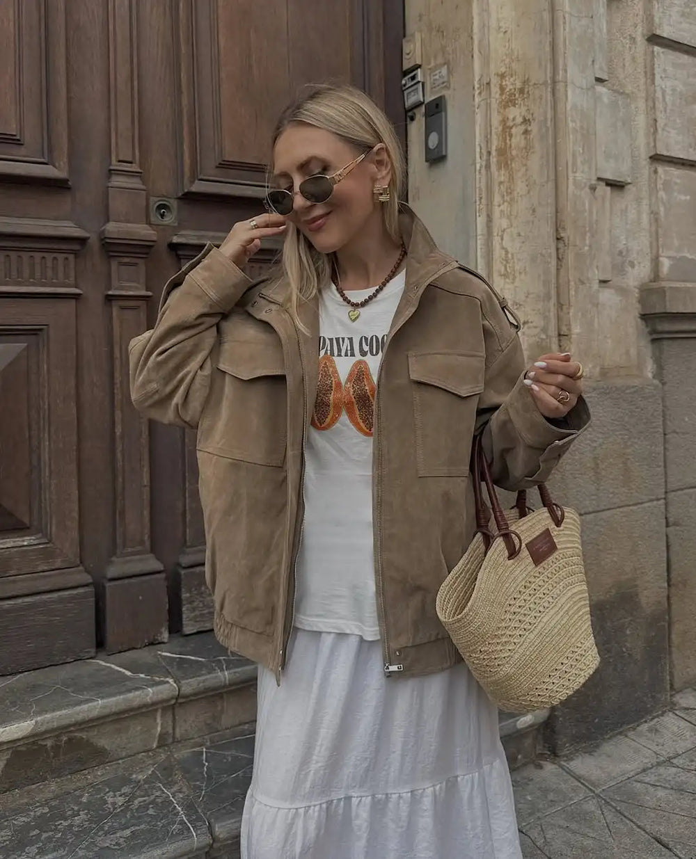 Woman in a beige jacket and white dress with a graphic shirt, holding a woven bag, standing in front of a wooden door.