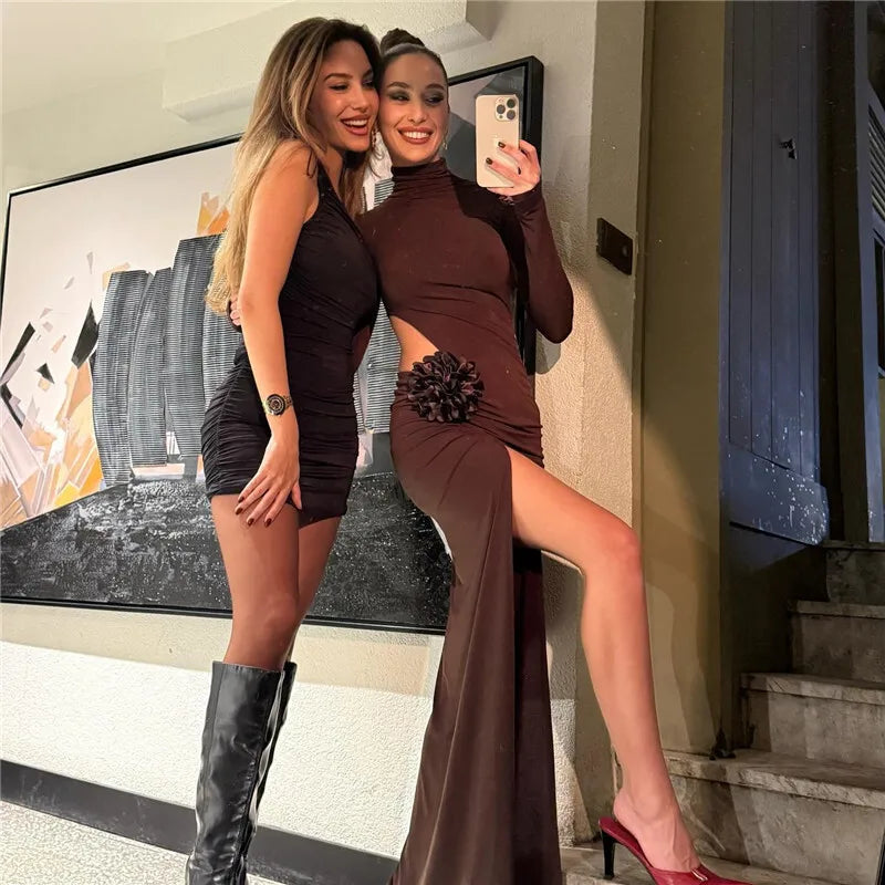 Two women taking a selfie in front of a painting, one in a black dress and the other in a brown dress with a high slit.