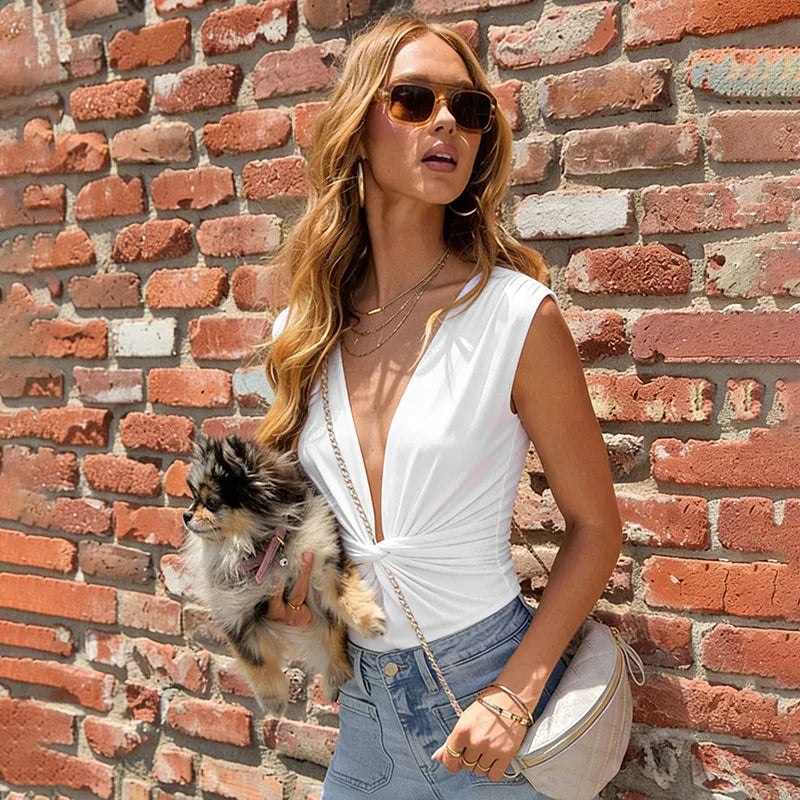 Woman in a white top and denim shorts standing against a brick wall with a small dog on her arm.