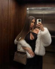 Woman taking a mirror selfie wearing a black dress and white fur coat in an elevator.