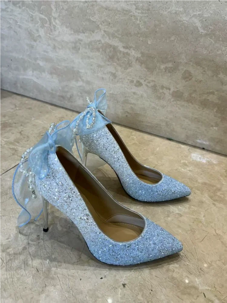 Pair of light blue glitter high heels with decorative ribbons on a beige floor.