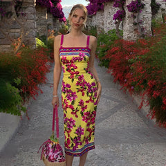 Woman in a yellow floral dress standing in a garden path with flowers and stone walls.