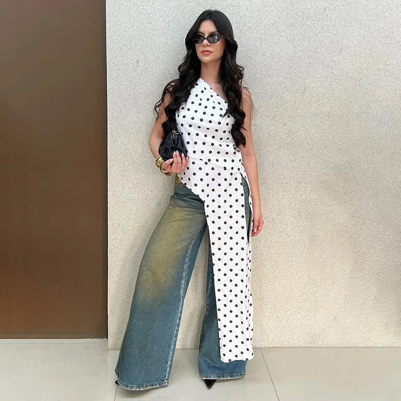 Woman wearing a white polka dot asymmetrical one-shoulder top with a long cascading hem and wide-leg jeans.