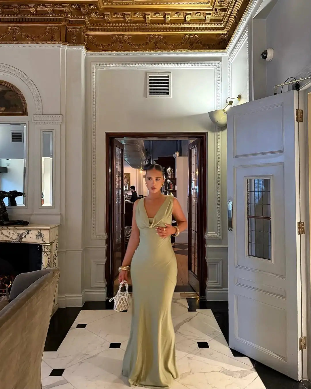 Woman in a light green evening gown standing in an elegant room with decorative ceiling and marble floor.