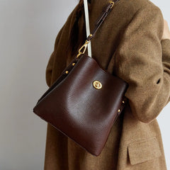 Brown leather handbag with gold accents worn over a brown coat on a neutral background