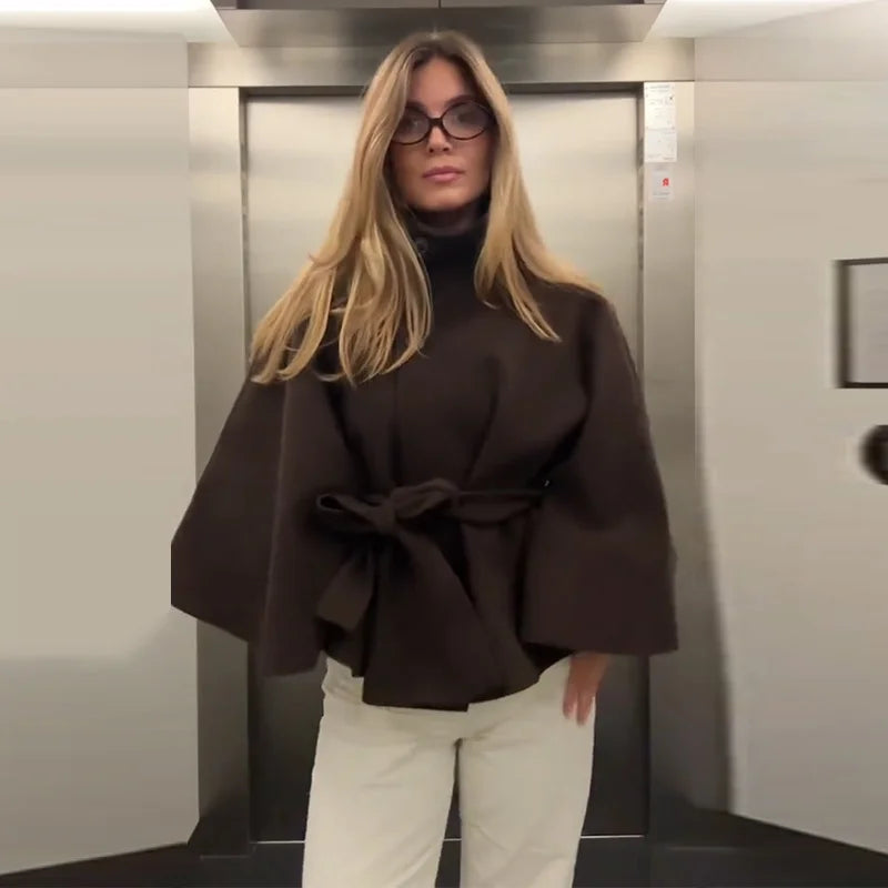 Woman wearing a brown cape and white pants in an elevator.