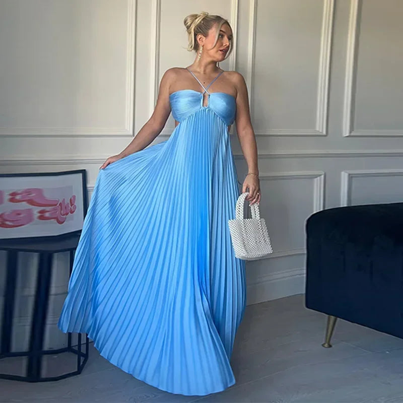 Woman in a blue pleated dress holding a white handbag in an indoor setting.