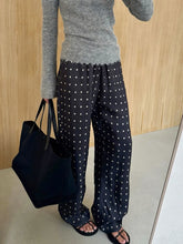 Person wearing a gray sweater and black polka dot pants holding a black bag against a wooden wall.