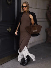 Woman in a brown dress with lace details and black coat holding a brown handbag against a dark door.