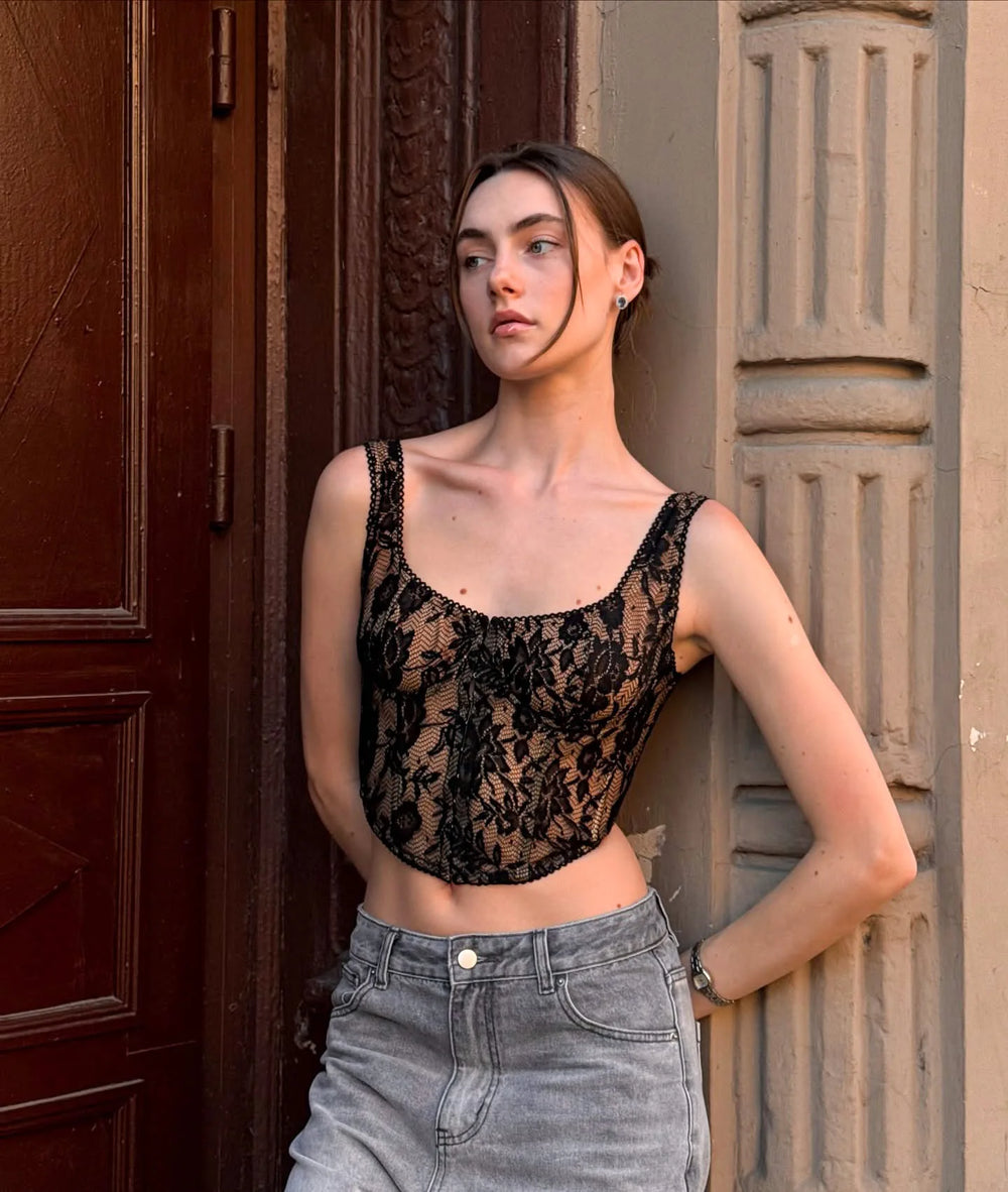 Woman wearing a black lace top and gray pants standing against a textured wall.