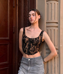 Woman wearing a black lace top and gray pants standing against a textured wall.