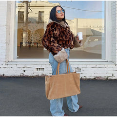 Person wearing a leopard print jacket and holding a brown bag, standing in front of a store window.