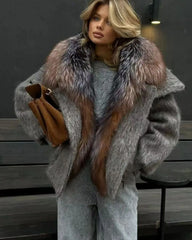Woman wearing a gray coat with a fur collar holding a brown bag against a dark background.