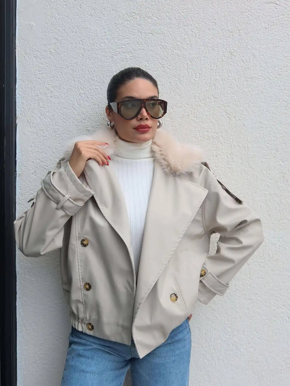 Woman wearing a beige trench coat with fur collar against a white wall