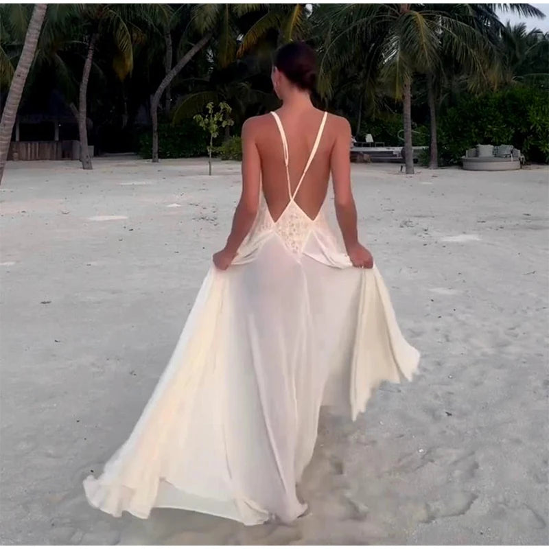 Woman in a white dress with a backless design walking on a sandy beach with palm trees in the background.