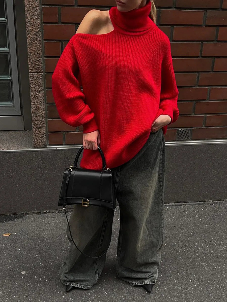 Person wearing a red sweater and dark jeans holding a black handbag against a brick wall.