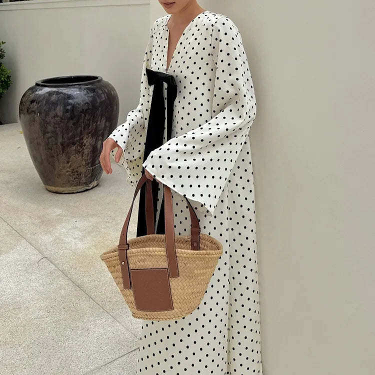 Person wearing a long white dress with black polka dots, holding a straw bag with brown leather straps.