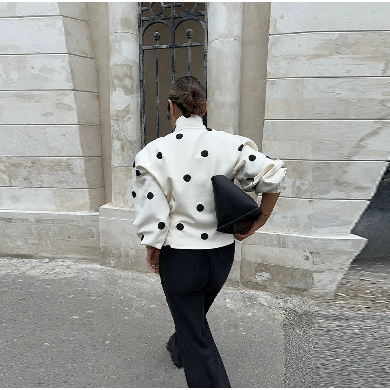 Person wearing a white polka dot jacket and black pants walking past a stone building.