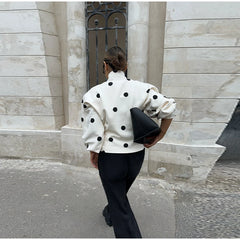 Person wearing a white polka dot jacket and black pants walking past a stone building.