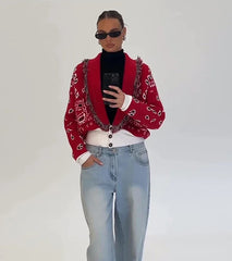 Person wearing a red patterned cardigan, black turtleneck, white shirt, and light blue jeans against a plain background.