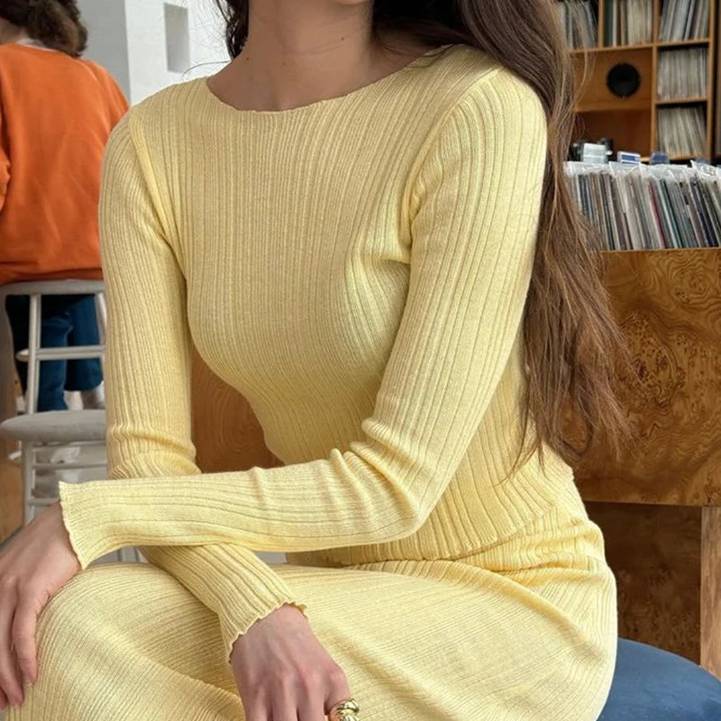 Person wearing a yellow ribbed dress sitting in a room with a wooden shelf in the background.