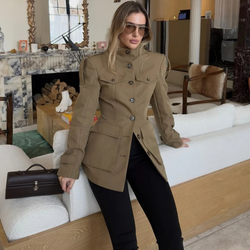 Woman in a beige coat standing in a living room with a fireplace and couch.