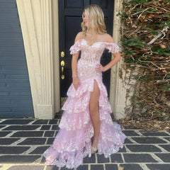 Woman in a pink off-shoulder dress with floral details standing outside a house.