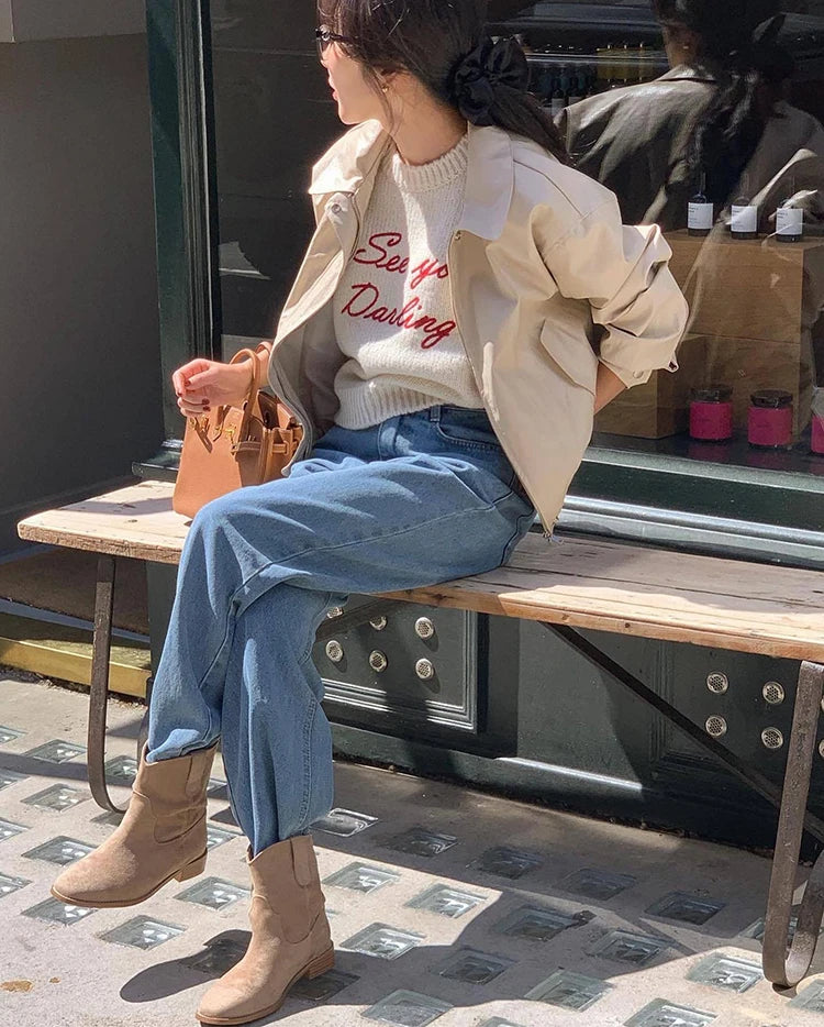 Person sitting on a bench wearing a beige jacket, white sweater with red text, blue jeans, and brown boots.