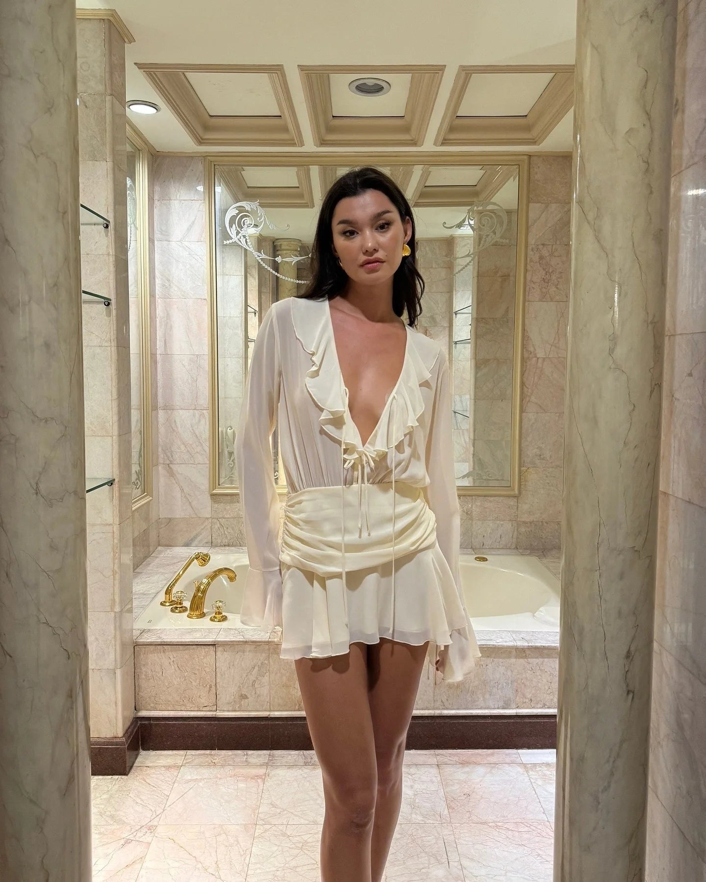 Woman in a white outfit standing in a bathroom with marble walls and a bathtub.
