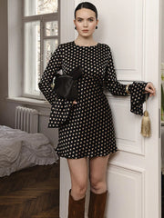 Woman in a black and white polka dot dress standing in a room with a window and bed in the background.
