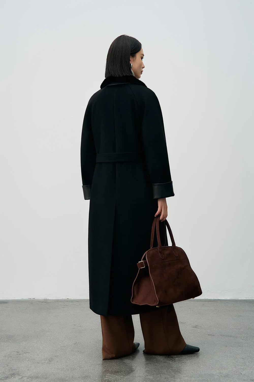 Person wearing a long black coat and brown pants holding a brown bag against a white background