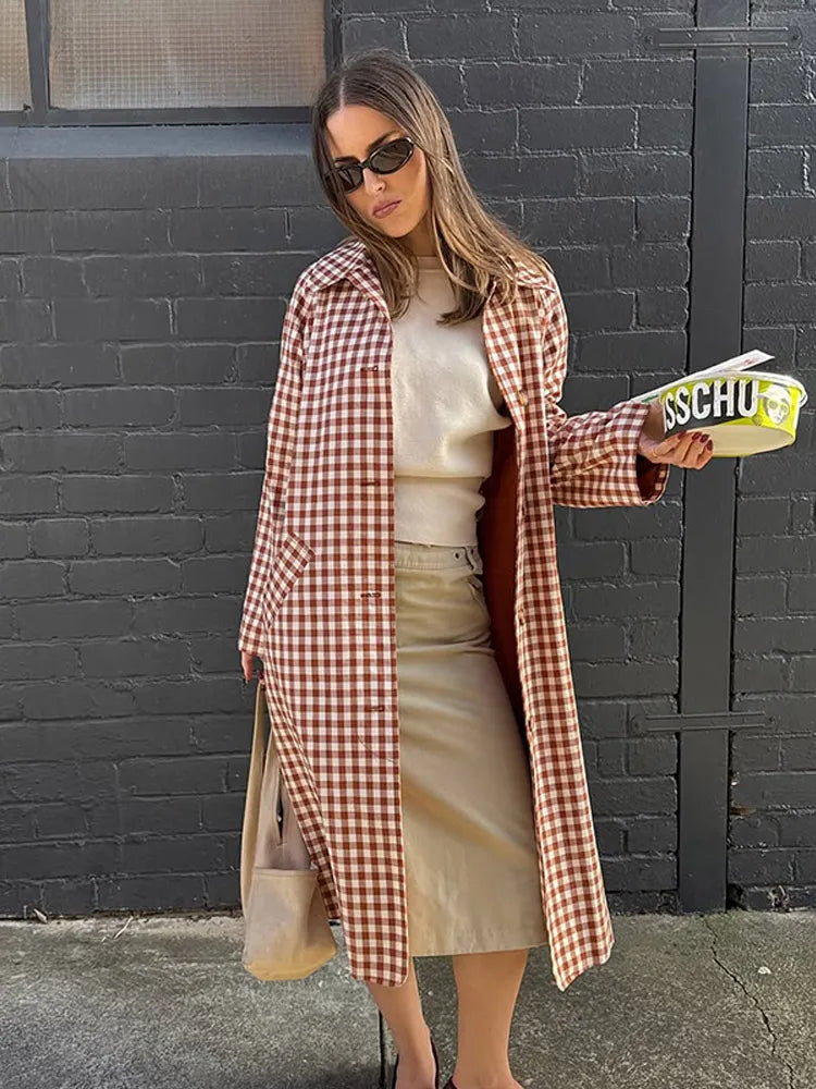 Woman wearing a checkered coat and sunglasses, holding a yellow shoe box against a black brick wall.