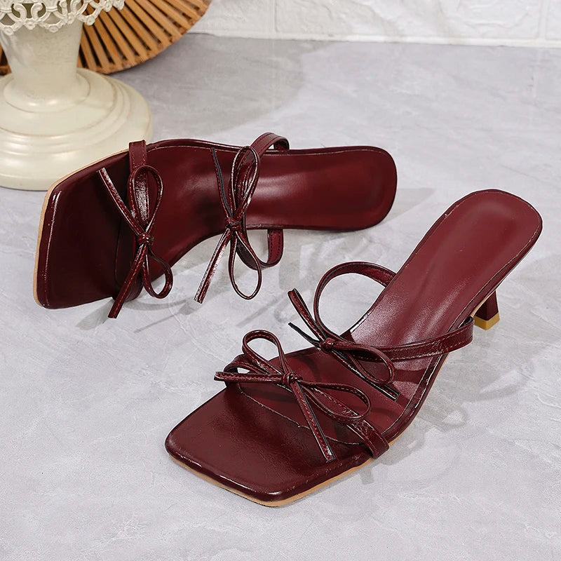 Pair of maroon high-heeled sandals with bow details on a light gray surface.