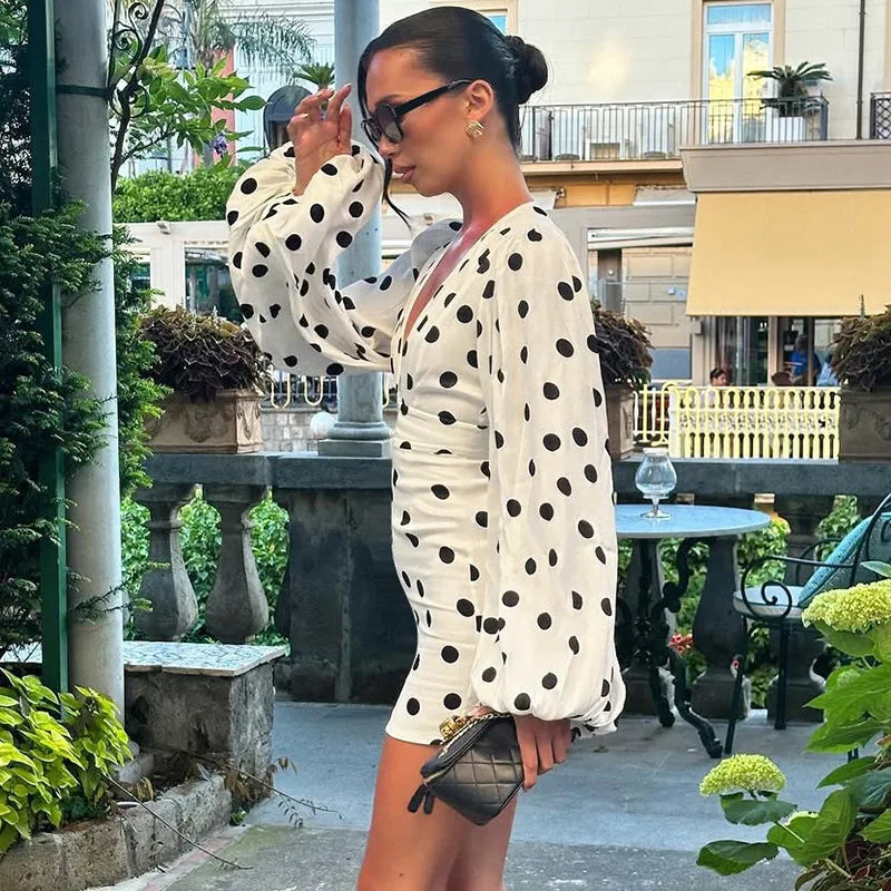 Woman in a polka dot dress standing outdoors with greenery and a building in the background