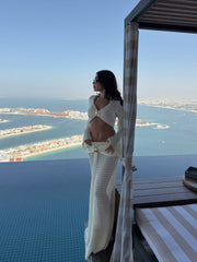 View of a woman in a white knit cropped long sleeve top and matching knit maxi skirt with draped detail, standing by a pool.