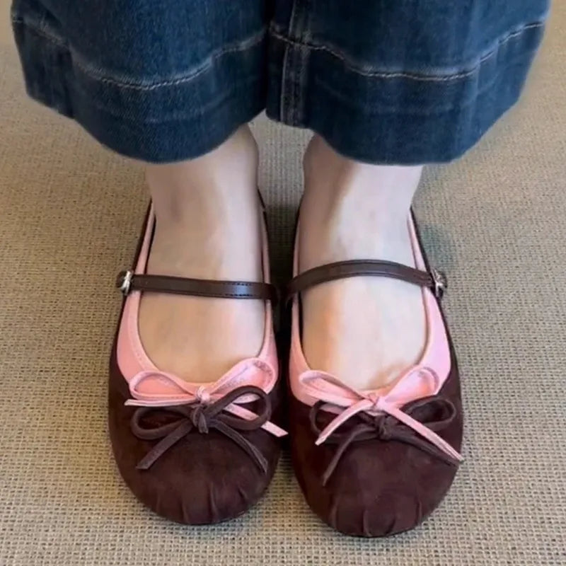 Brown ballet flats with pink ribbons worn by a person on a textured surface.