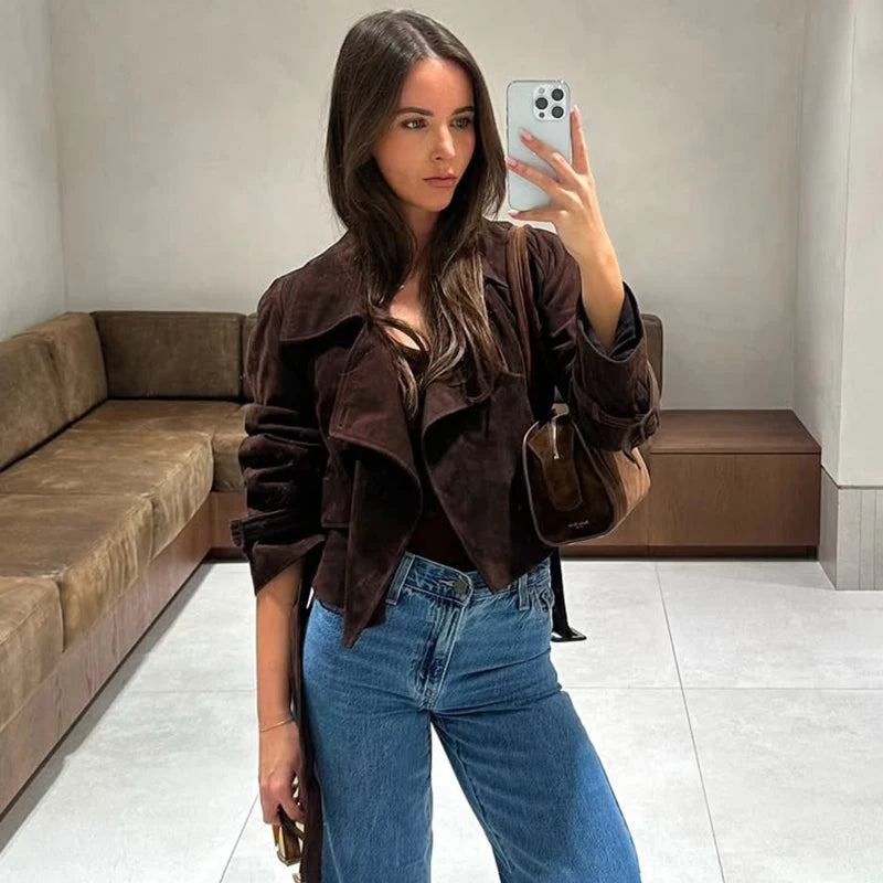 Woman taking a mirror selfie wearing a brown jacket and blue jeans in a room with a brown couch.