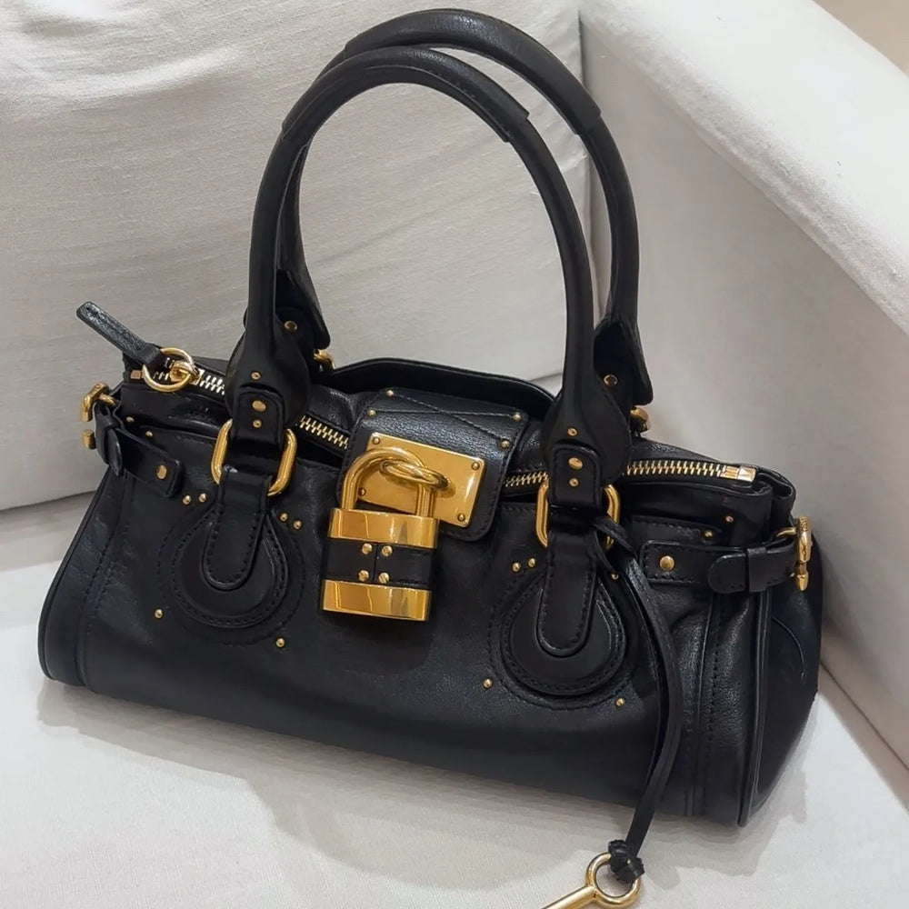 Black leather tote bag with gold lock closure on a white couch.